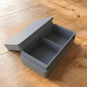 Könnte beinhalten: Eine graue rechteckige Box mit einem Klappdeckel, unterteilt in zwei Fächer. Die Box ist geöffnet und gibt den Innenraum frei. Die Box steht auf einer Holzoberfläche.