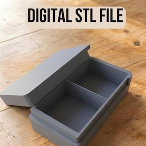 May include: A gray rectangular box with a hinged lid, divided into two compartments. The box is open, revealing the interior. The text "DIGITAL STL FILE" is at the top. The box is on a wooden surface.
