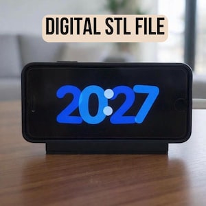May include: A black smartphone displaying the time 20:27 in blue digits, resting on a black charging stand. The phone is connected to a white charging cable. The text "DIGITAL STL FILE" is in a beige rectangle above the phone.