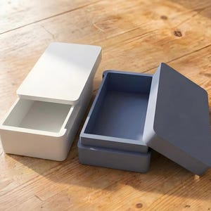 Könnte beinhalten: Zwei rechteckige Boxen mit Deckeln, eine weiße und eine blau-graue, liegen auf einer Holzoberfläche. Die weiße Box hat ihren Deckel leicht geöffnet, wodurch das Innere sichtbar wird. Die blau-graue Box ist offen.