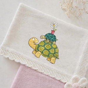 Può includere: Una coperta per neonati in maglia color crema con un bordo smerlato. La coperta presenta un disegno ricamato di due tartarughe, una che trasporta l'altra, con un fiore in un vaso. Una coperta rosa è parzialmente visibile sotto.