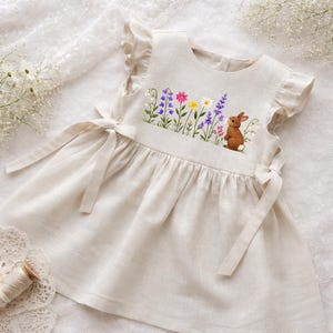 May include: A cream-colored baby dress with embroidered flowers and a brown bunny. The dress features ruffled sleeves and tie closures at the sides. The embroidered flowers include purple, pink, yellow, and white blooms.