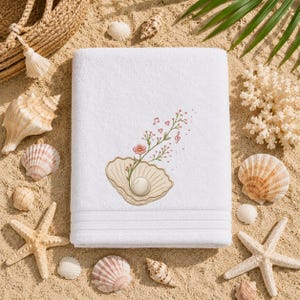 Puede incluir: Una toalla de baño rectangular blanca con un diseño bordado de una concha marina abierta que contiene una perla, con un tallo verde delicado, pequeñas flores rosas, corazones rojos diminutos y notas musicales que emergen de la concha.