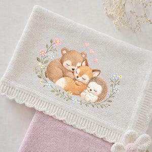 Puede incluir: Una manta para bebé de color gris claro con un diseño bordado de un oso, un zorro y un erizo acurrucados juntos, rodeados por una corona floral. La manta tiene un borde festoneado y está superpuesta a una manta rosa.