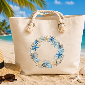 Puede incluir: Un bolso tote de lona beige con asas de cuerda y un diseño bordado circular de conchas marinas y estrellas de mar en tonos azules y beige. El bolso está en una playa de arena con un sombrero de paja y gafas de sol.