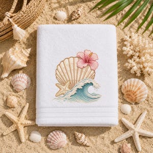 May include: A white rectangular bath towel featuring an embroidered design of a large tan seashell, a pink hibiscus flower, a small tan starfish, and a blue ocean wave. The towel has a textured border with three horizontal lines near the bottom.