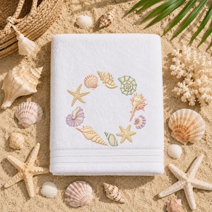 May include: White beach towel with an embroidered seashell wreath design. The wreath features various colorful seashells and starfish. The towel is surrounded by seashells, starfish, and sand, creating a coastal theme.