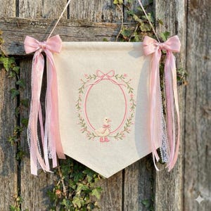 May include: A cream-colored banner with a pink duck illustration, surrounded by a floral wreath and oval frame. Pink bows and ribbons adorn the top corners. The banner hangs on a wooden door.