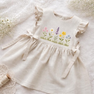 May include: A cream-colored linen dress with embroidered flowers in pink, white, yellow, purple, and blue. The dress features ruffled sleeves and tie closures at the sides. The floral embroidery is above the skirt, which has a gathered waist.