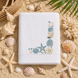 May include: A white rectangular bath towel featuring an embroidered L-shaped border of blue and beige seashells, starfish, and sand dollars. The towel has a textured fabric surface with three horizontal lines embossed near the bottom edge.