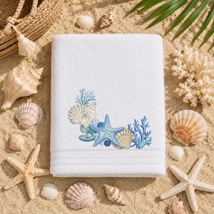 May include: White beach towel with embroidered seashells, starfish, and coral in shades of blue and beige. The towel is surrounded by seashells and starfish on a sandy surface, evoking a coastal theme.