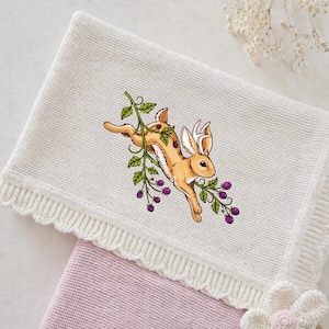 Può includere: Una coperta per neonati in maglia color crema con bordo smerlato. La coperta presenta un'illustrazione ricamata di un coniglio marrone che salta attraverso un ramo di bacche. Sotto è visibile una seconda coperta rosa.