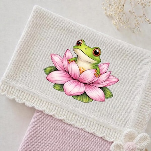 May include: A white knitted baby blanket with a decorative edge. The blanket features a colourful illustration of a green frog sitting in a pink lotus flower. The blanket is folded over a pink blanket.