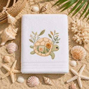 May include: A white rectangular towel featuring an embroidered design of a light green turtle with a tan shell. The shell contains floral and botanical elements in shades of orange, pink, and green. The towel has a textured border at the bottom.
