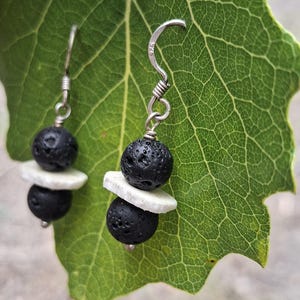 May include: A pair of dangle earrings featuring two porous black lava beads separated by a flat white disc. The beads are suspended from silver-toned metal hooks, with the metal hook on the right earring clearly stamped with the number 925.