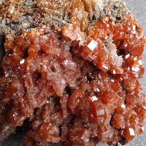 May include: A close-up of a mineral specimen with striking orange-red crystals. The crystals are geometrically shaped and clustered on a darker, earthy matrix. The overall appearance is rough and natural, with a sparkling texture.