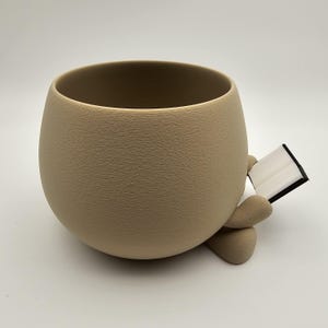 May include: A beige ceramic planter with a textured surface, shaped like a rounded cup. The planter has two small, rounded feet and is holding a small, open book with white pages and a black cover. The planter is on a white background.