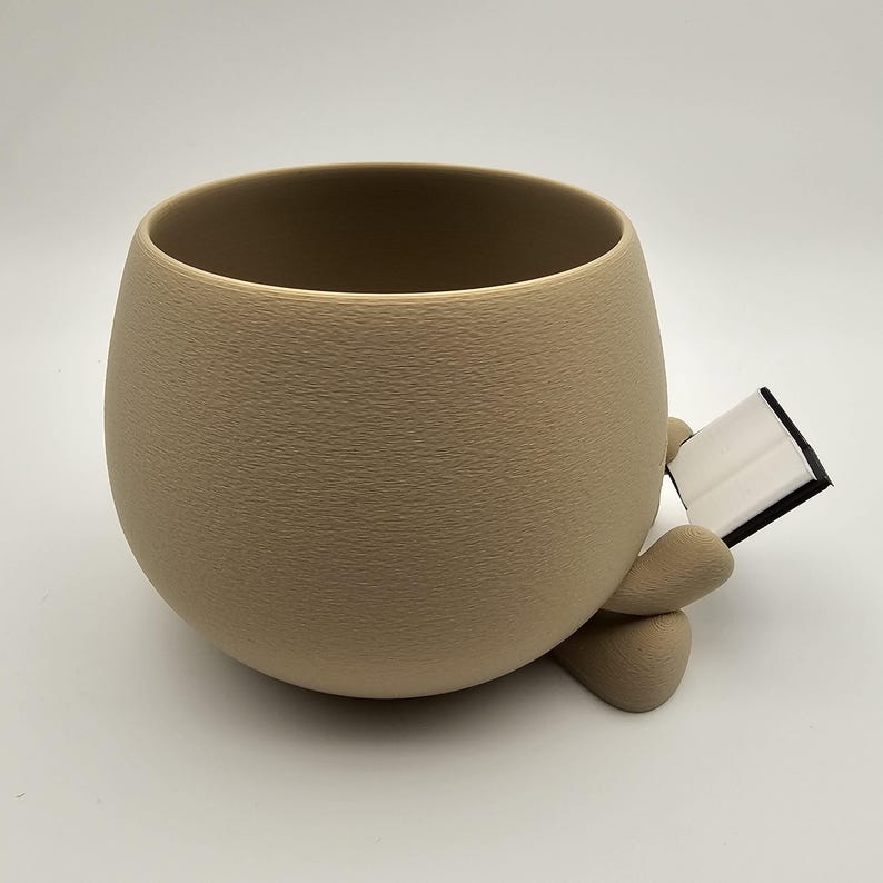 May include: A beige ceramic planter with a textured surface, shaped like a rounded cup. The planter has two small, rounded feet and is holding a small, open book with white pages and a black cover. The planter is on a white background.