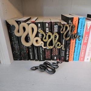 Puede incluir: Cuatro figuras decorativas con forma de serpiente en diferentes colores y acabados. Una es beige mate, una es dorada metálica, una es bronce metálico oscuro y una es gris carbón oscuro. Las serpientes están colocadas sobre y frente a libros.