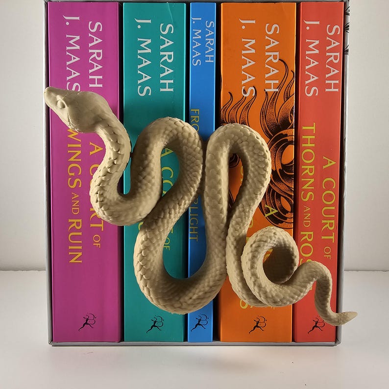 May include: A beige, textured snake bookend coiled around a row of colorful books. The books have titles by Sarah J. Maas. The spines are pink, teal, blue, orange, and red. The bookend adds a decorative touch to a bookshelf.
