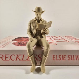 May include: A tan figurine of a person in a cowboy hat, shirt, and boots, seated and reading a book. The figurine is sitting on a book with the title "Reckless" and the author's name "Elsie Silver" visible.