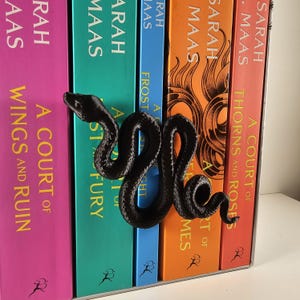 May include: A black, 3D-printed snake bookmark rests on a row of colorful books. The books have titles like "A Court of Thorns and Roses" and "A Court of Wings and Ruin" by Sarah Maas. The spines are in shades of pink, teal, blue, orange, and red.