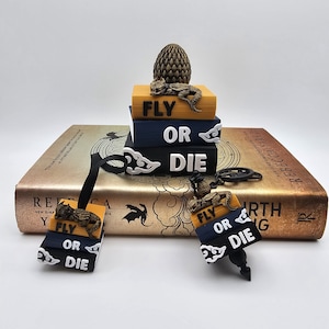 May include: A stack of three miniature books with yellow, blue, and black spines featuring the text FLY OR DIE, topped with a textured dragon egg and a small dragon figure. Two matching keychains with similar book designs and dragon figures are included.