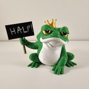 May include: A green frog figurine wearing a gold crown and holding a sign that reads "HALP!". The frog has a white belly and yellow eyes. The sign is black with white lettering.