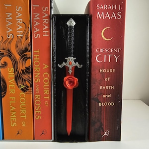 May include: A decorative sword bookend with a red blade and a black handle, adorned with a silver hilt and a red rose. The bookend is placed between several books with titles by Sarah J. Maas.