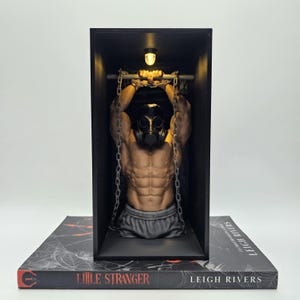 May include: A muscular torso figurine wearing a black gas mask, with wrists shackled to a horizontal bar by metal chains, housed inside a black rectangular display box. The base is a book titled Little Stranger by Leigh Rivers.