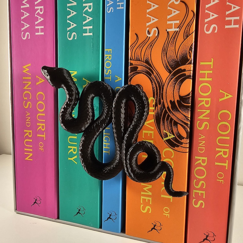 May include: A black, serpentine-shaped bookend rests against a row of colorful books. The books have titles like "A Court of Wings and Ruin" and "Thorns and Roses." The bookend adds a decorative touch to the bookshelf.