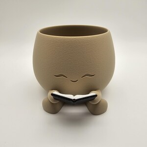 May include: A tan-colored ceramic planter with a smiling face design. The planter is holding a small black and white book. The planter has a textured surface and a rounded shape, perfect for small plants or succulents.