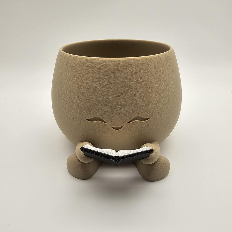 May include: A tan-colored ceramic planter with a smiling face design. The planter is holding a small black and white book. The planter has a textured surface and a rounded shape, perfect for small plants or succulents.