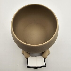 May include: A beige ceramic planter with a round bowl shape. The planter has two small arms holding a book with white pages and a black cover. The planter is designed to hold plants.