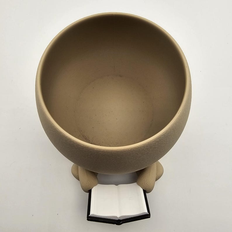 May include: A beige ceramic planter with a round bowl shape. The planter has two small arms holding a book with white pages and a black cover. The planter is designed to hold plants.