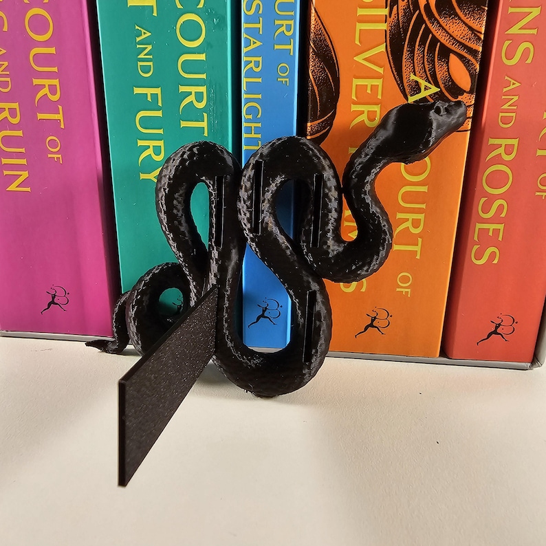 May include: A black, 3D-printed snake-shaped bookend. The bookend is positioned in front of a row of colorful books with titles including "Court of Silver Flames" and "A Court of Thorns and Roses".