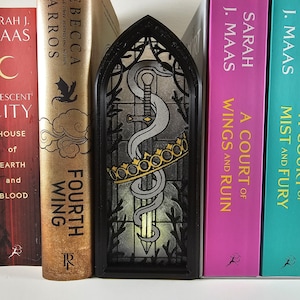 Könnte beinhalten: Schwarzer dekorativer Buchstütze mit einem Buntglasdesign, das ein Schwert, eine Krone und eine Schlange zeigt. Die Buchstütze befindet sich zwischen Büchern, darunter Titel wie "A Court of Wings and Ruin" und "Fourth Wing".
