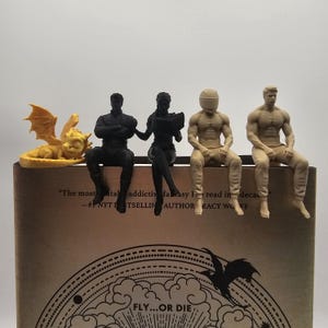 May include: A collection of five small 3D printed figurines perched on the edge of a book. The set includes a yellow dragon, two black figures, and two beige figures. The book cover features the text "The most addictive fantasy I've read in decades" and "FLY...OR DIE".