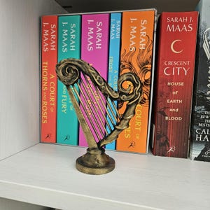 May include: A small, decorative harp figurine with a metallic gold and black finish, featuring a curved frame with intricate scrollwork details and thin vertical strings, standing on a circular base in front of a row of colorful hardcover books.