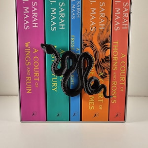 May include: A black snake-shaped bookend rests against a row of colorful books. The books have titles like "A Court of Thorns and Roses" and "A Court of Wings and Ruin" by Sarah J. Maas. The bookend adds a decorative touch.