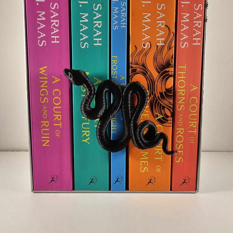 May include: A black snake-shaped bookend rests against a row of colorful books. The books have titles like "A Court of Thorns and Roses" and "A Court of Wings and Ruin" by Sarah J. Maas. The bookend adds a decorative touch.