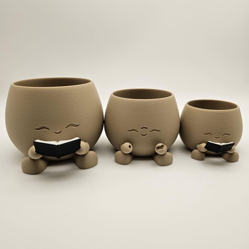May include: Three beige ceramic planters with smiling faces and small black books. The planters have rounded bodies, small legs, and open tops for plants. The smallest planter is on the right, the largest on the left.
