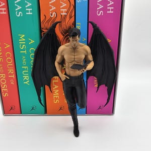 May include: A figurine of a shirtless male figure with large black wings, wearing black pants and holding a small black book. The figure has a muscular build and dark hair, standing in front of a row of colorful books with visible text.