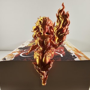 May include: A golden-colored, 3D-printed figurine of a woman with flowing hair and a flame held in her hand. The figurine is perched on top of a book. The woman's form is detailed with intricate patterns, and the flame is a vibrant orange.