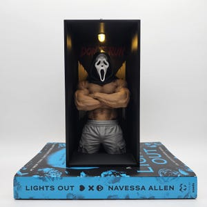 May include: A muscular Ghostface figure wearing a black hood and grey shorts, displayed inside a black rectangular box with an overhead light. The box sits on a blue book titled LIGHT OUT by Navessa Allen, featuring the text DON'T RUN in the background.