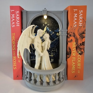 May include: A gray book nook featuring a balcony with a winged figure and a person standing together. The background shows a dark mountain landscape with a starry sky and a small light at the top. The book nook is placed between two books by Sarah J. Maas.