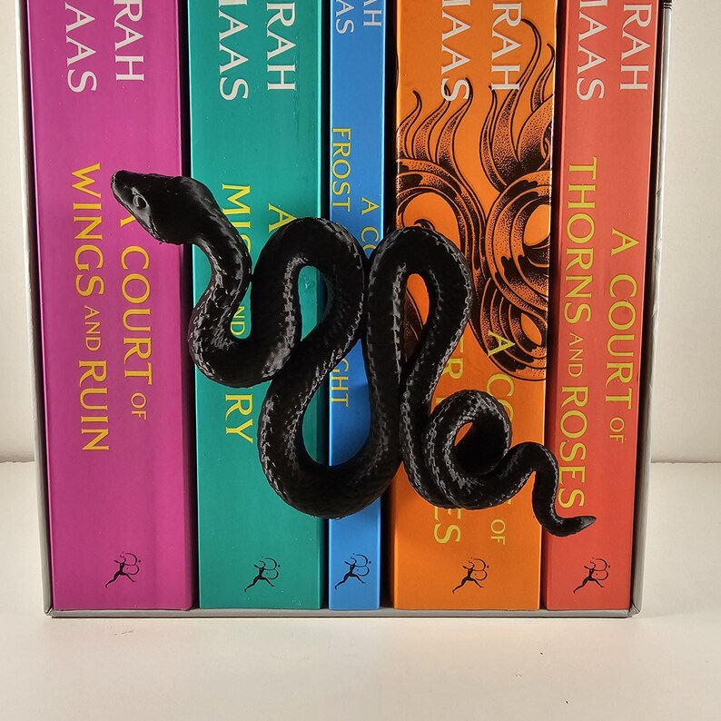 May include: A black, serpentine-shaped bookend rests against a row of colorful books. The bookend is a detailed, three-dimensional design. The books have titles like "A Court of Thorns and Roses" and "Wings and Ruin."