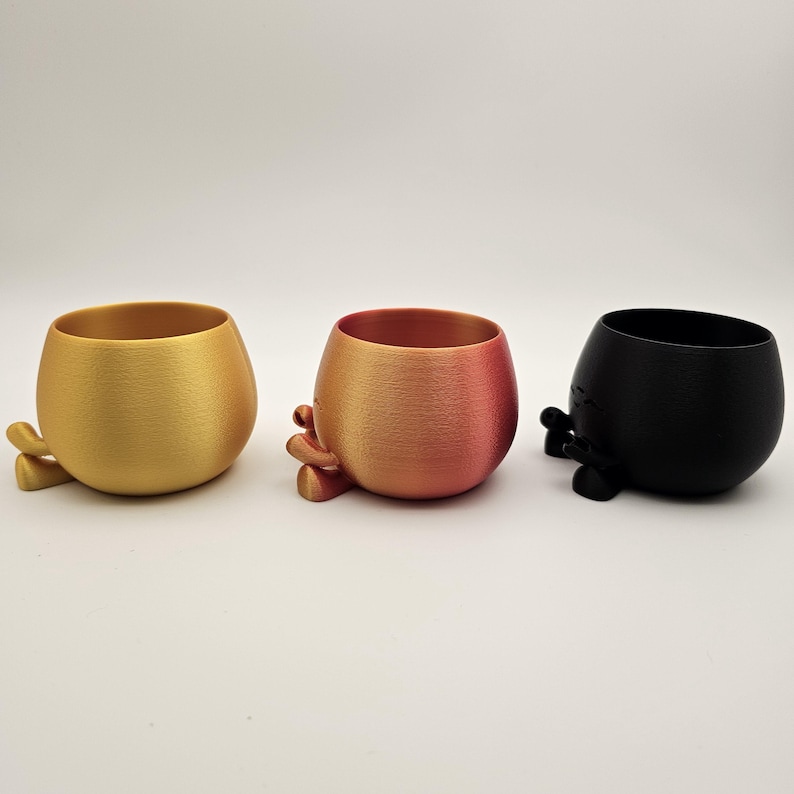 May include: Three small, rounded planters in gold, orange-red, and black. Each planter has a textured surface and small, stylized feet. The gold planter is on the left, the orange-red in the middle, and the black on the right.