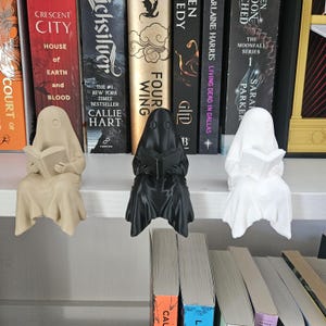 May include: Three small ghost figurines sitting on a shelf edge, each holding an open book. The ghosts are colored beige, black, and white, featuring smooth, draped fabric-like textures and simple circular eye cutouts on their hooded heads.
