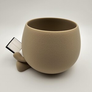 May include: A beige, textured ceramic planter with a rounded shape. The planter has a wide opening and a small, attached support structure. The planter is empty and ready for plants. The background is white.